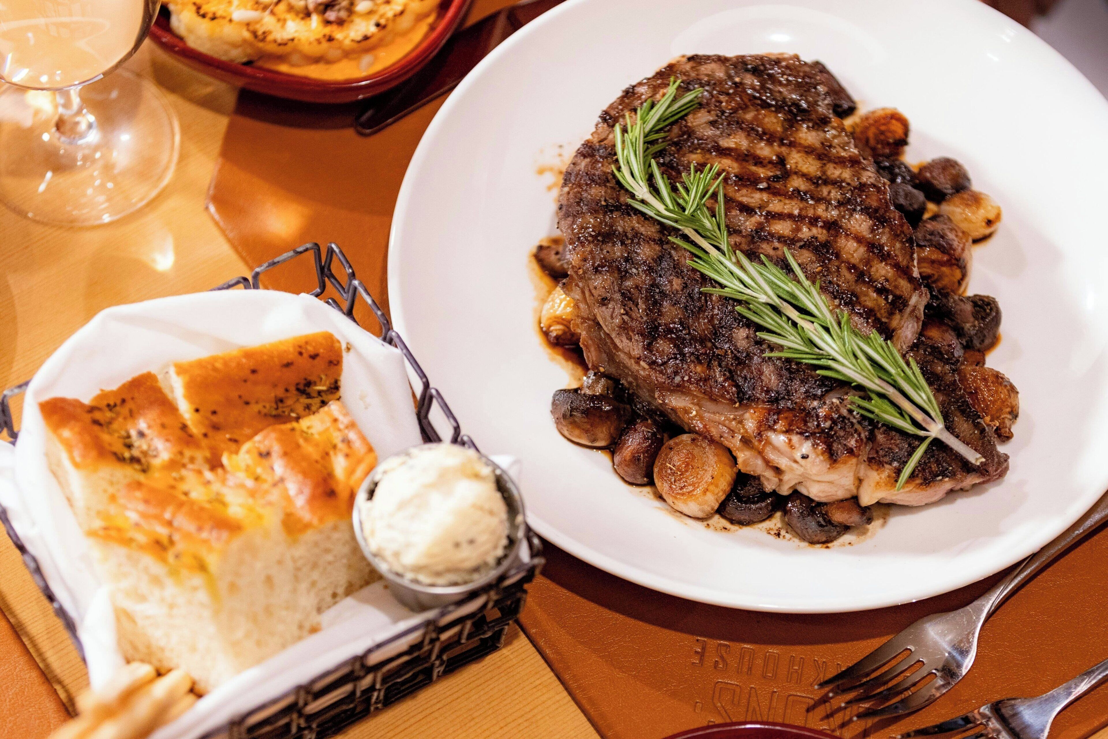 A plate of grilled steak with mushrooms and rosemary, accompanied by a basket of bread and a small bowl of butter.
