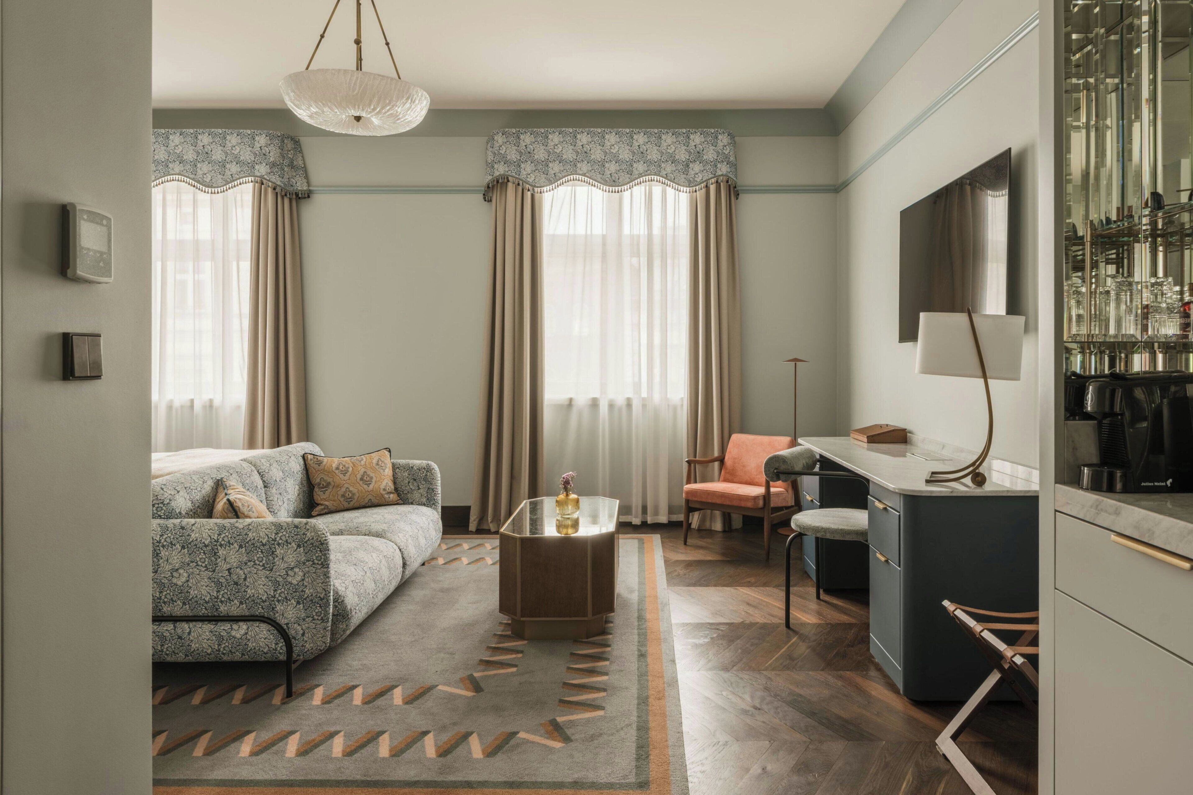Habitación de hotel moderna con sofá, sillón, mesa de centro y una zona de cocina con espejo y electrodomésticos.