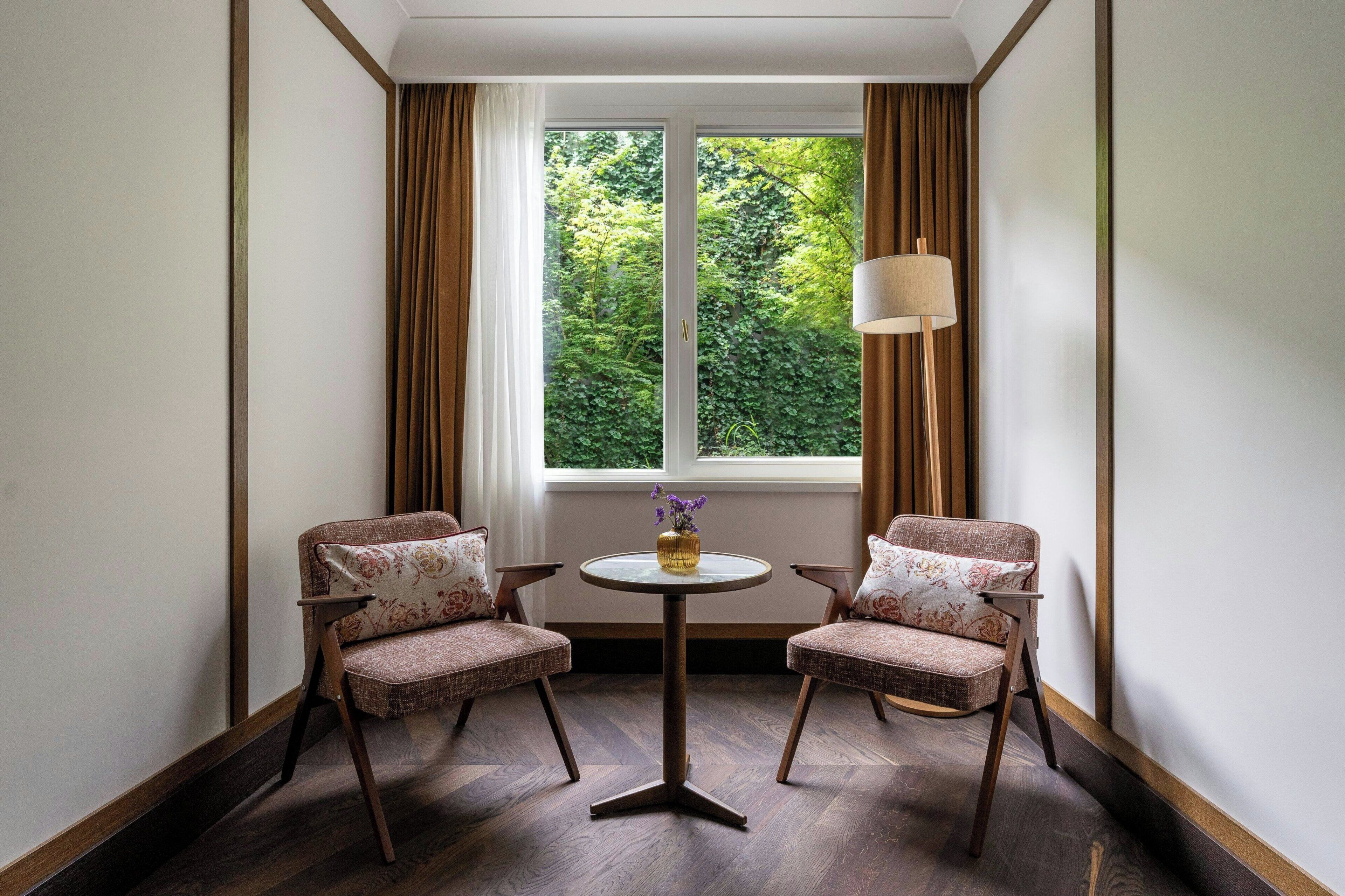 A corner room with two upholstered chairs, a small round table, and a lamp. A window with curtains and a view of greenery is visible.
