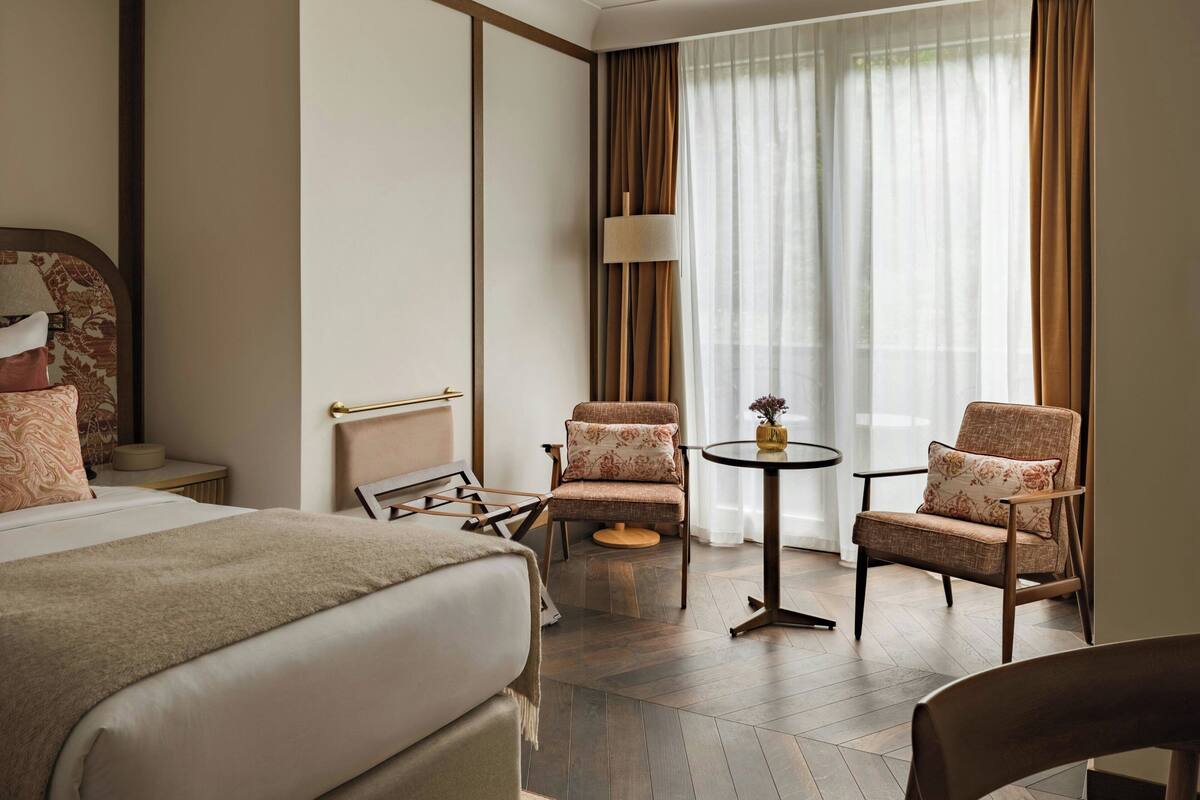 Ein Hotelzimmer mit einem Bett, zwei Sesseln, einem kleinen runden Tisch, einer Lampe und einem großen Fenster mit Vorhängen.