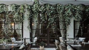 Un área de comedor al aire libre con mesas y sillas, rodeada de amplias ventanas y cubierta por un dosel de hiedra.