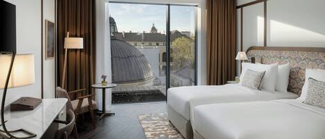 A hotel room with two beds, a desk with a chair, a small table, and a view of the cityscape through large windows.