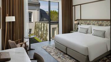 A modern hotel room with a large bed, a desk with a laptop, a small table, and a view of a garden through a sliding door.