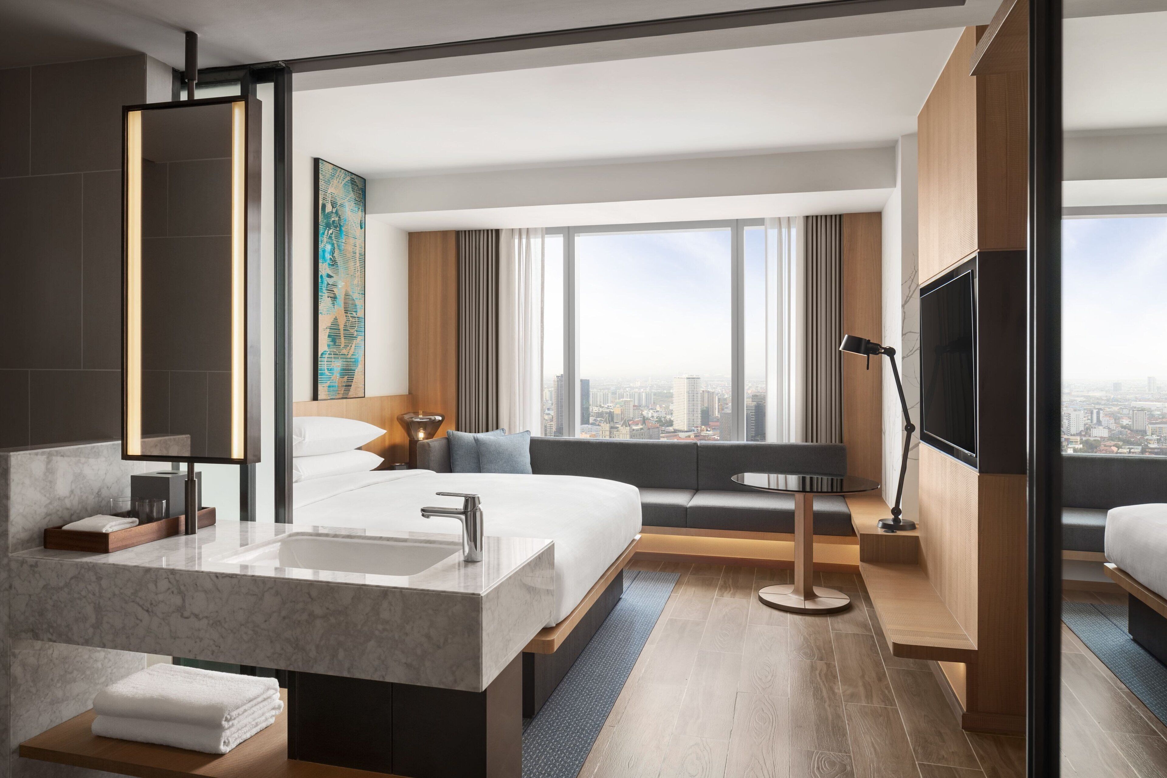 A modern hotel room with a large bed, a bathroom with a marble vanity, a large window with a city view, and a flat-screen TV.