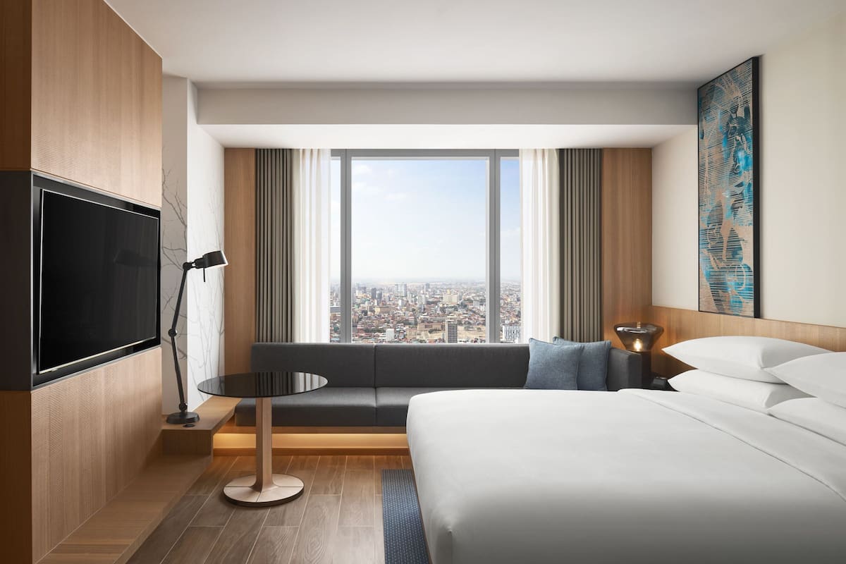 A hotel room with a bed, a sofa, a TV, a lamp, and a large window with a city view.
