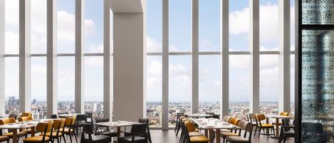 A spacious dining area with large windows offering a city view, modern furniture, and a clean, minimalist design.