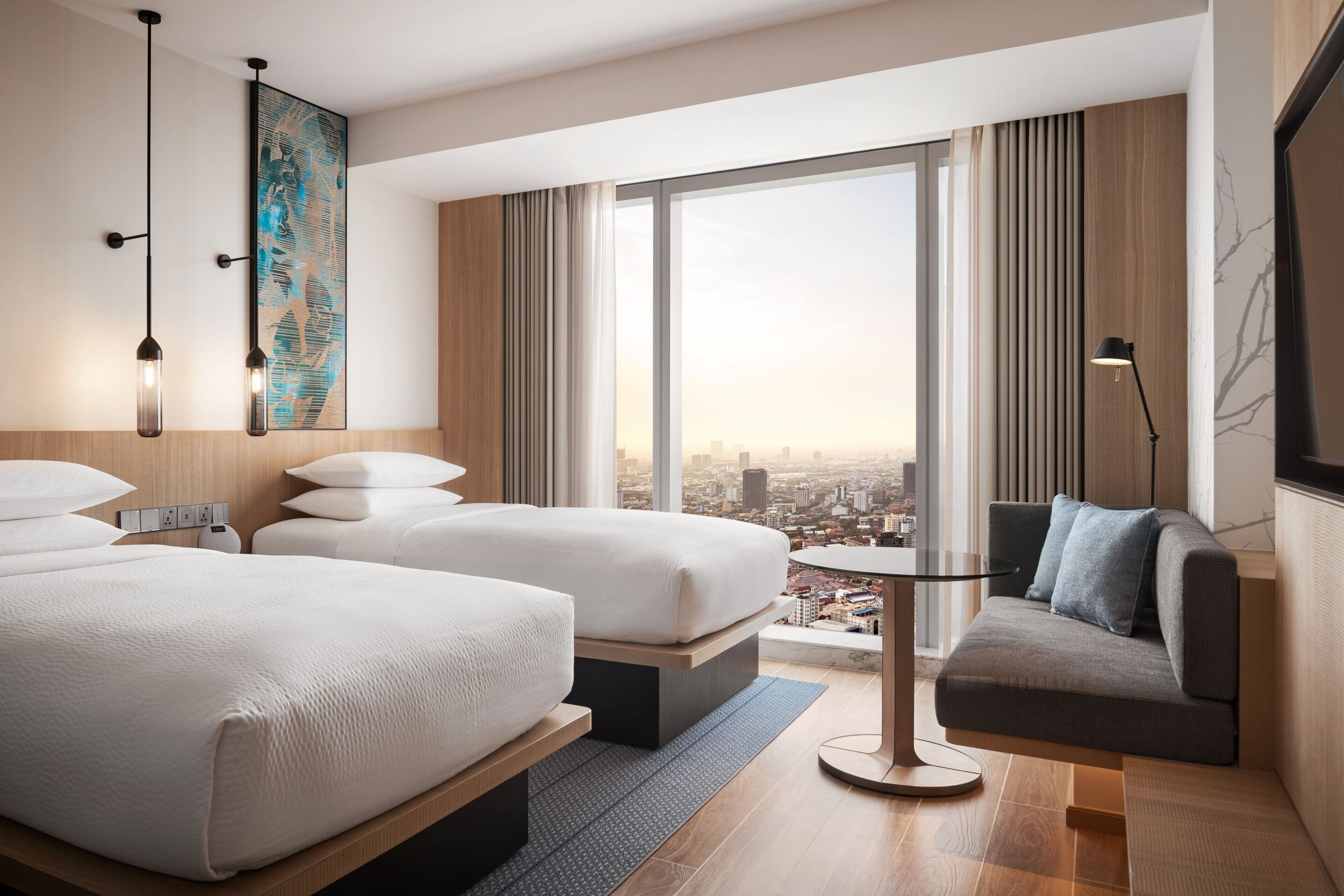 A modern hotel room with two beds, a sofa, a small table, and a city view.