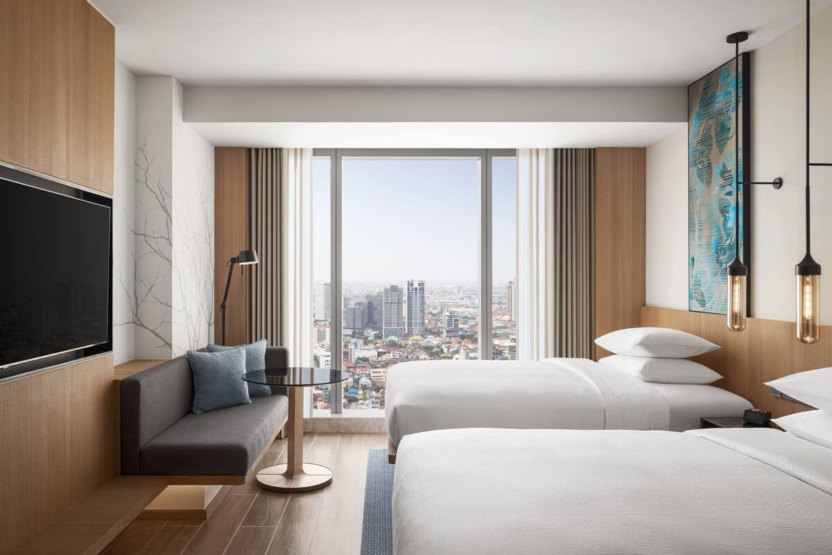 A modern hotel room with a city view, a sofa, two beds, and a flat-screen TV.