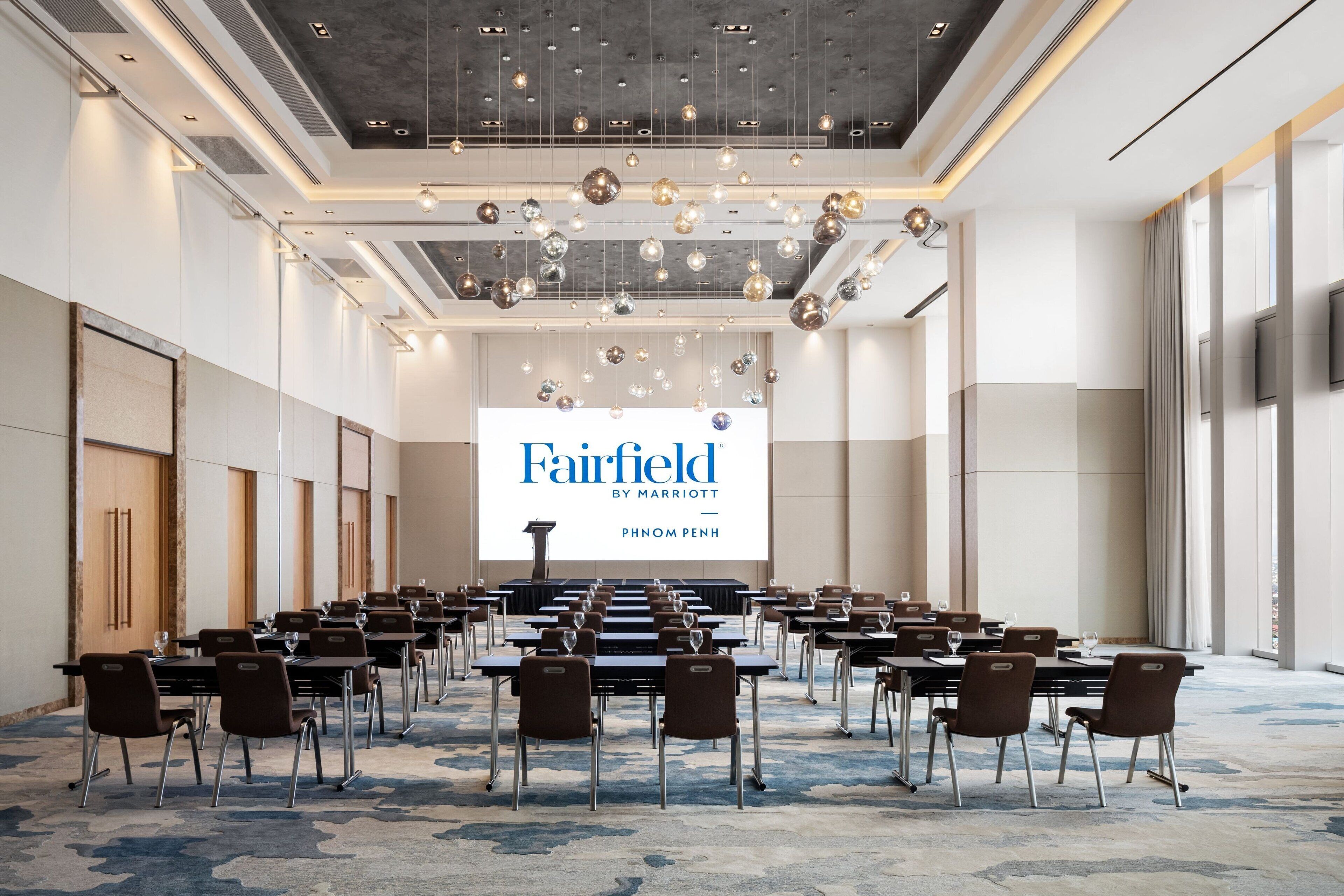 A conference room with a large screen displaying "Fairfield by Marriott" and "Phnom Penh". The room has rows of tables and chairs, a patterned carpet, and a modern ceiling with hanging lights.