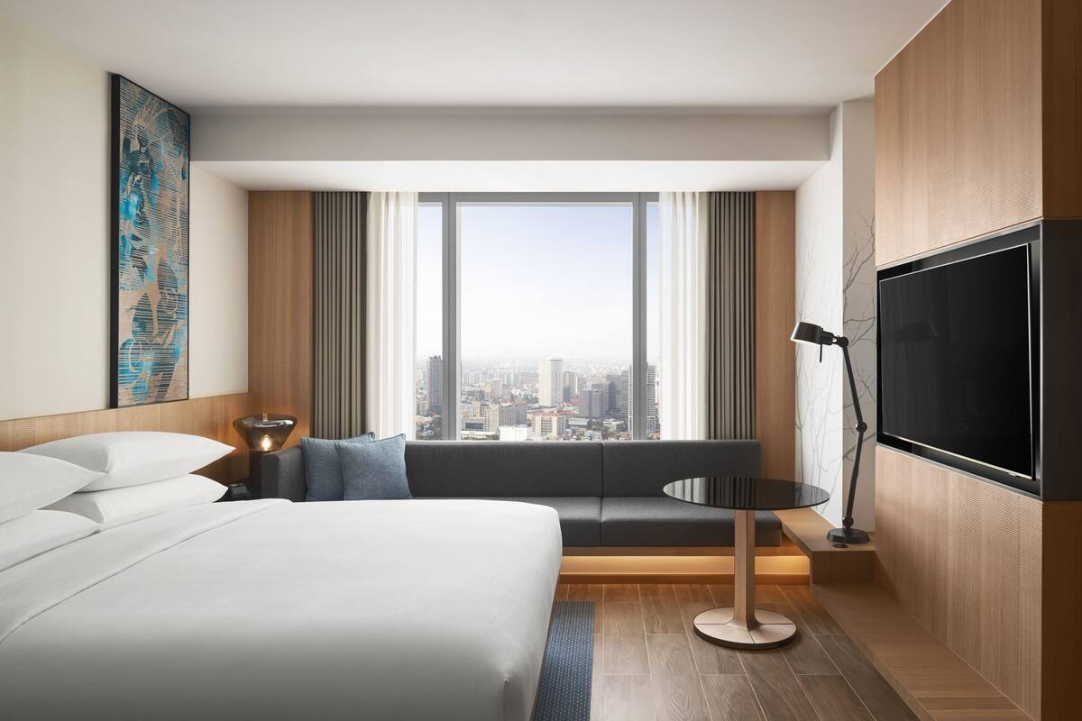 A modern hotel room with a large bed, a seating area with a view of the city, a flat-screen TV, and a large window with curtains.