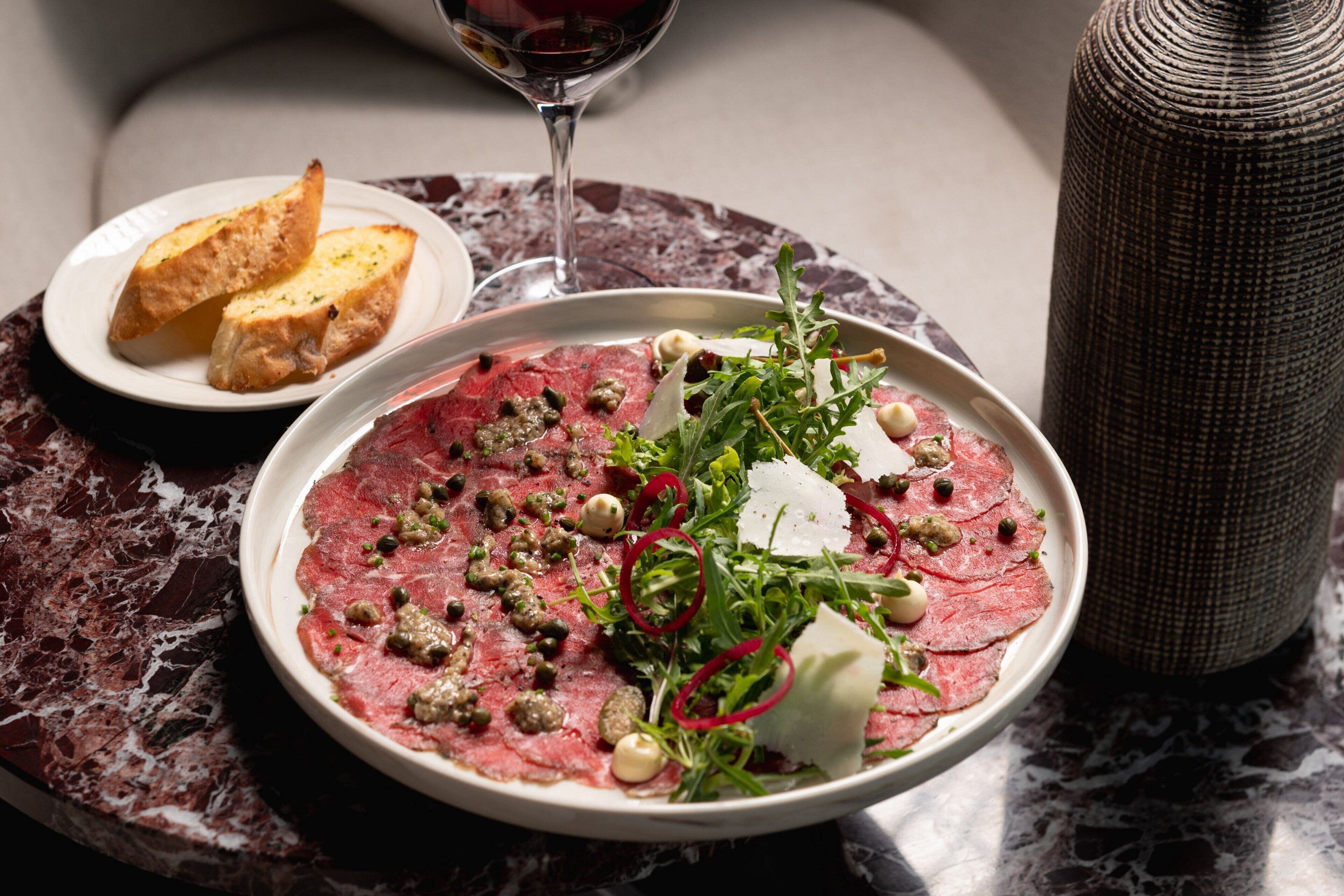 A plate of steak tartare with capers and greens, a glass of red wine, and toasted bread on a marble table.