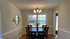 A dining room with a round table, four chairs, a chandelier, and a large window with blinds. - 2.0 to the Masters! (Augusta)