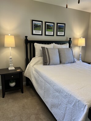 Una cama bien hecha con edredón blanco y dos almohadas, flanqueada por dos mesitas de noche con lámparas, frente a una pared con tres cuadros enmarcados.