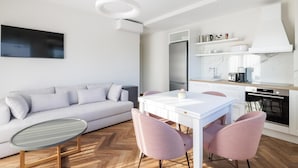 A modern living room with a white sofa, a glass coffee table, and pink chairs. A kitchen with white cabinets and a built-in oven is visible. - Deluxe Apartment (Thasos)