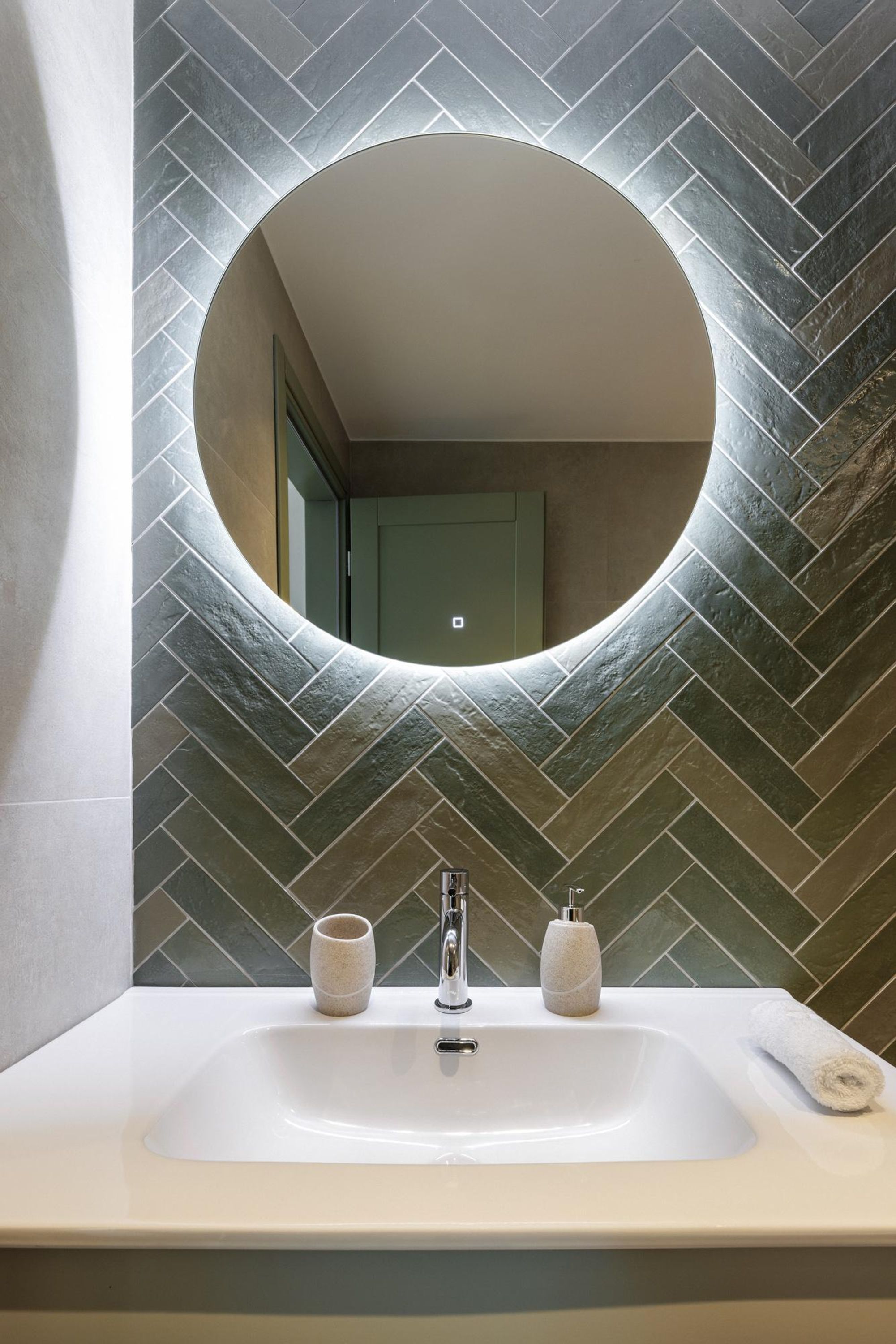 A bathroom with a white sink, two soap dispensers, and a round mirror with a herringbone tile backsplash.