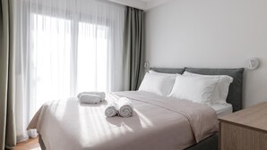 A neatly made bed with white linens and two rolled towels, a wooden nightstand, and a window with sheer curtains. - Deluxe Apartment (Thasos)