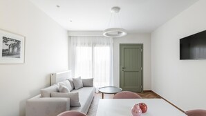 A modern living room with a sofa, a dining table with chairs, a television mounted on the wall, and a door. - Premium Apartment (Thasos)