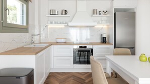 A modern kitchen with white cabinetry, a built-in oven, a stainless steel refrigerator, and a window with a curtain. - Superior Apartment (Thasos)