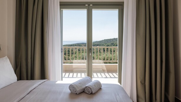 A hotel room with a bed, two rolled towels, and a balcony with a view of greenery and a distant landscape. - Superior Apartment (Thasos)