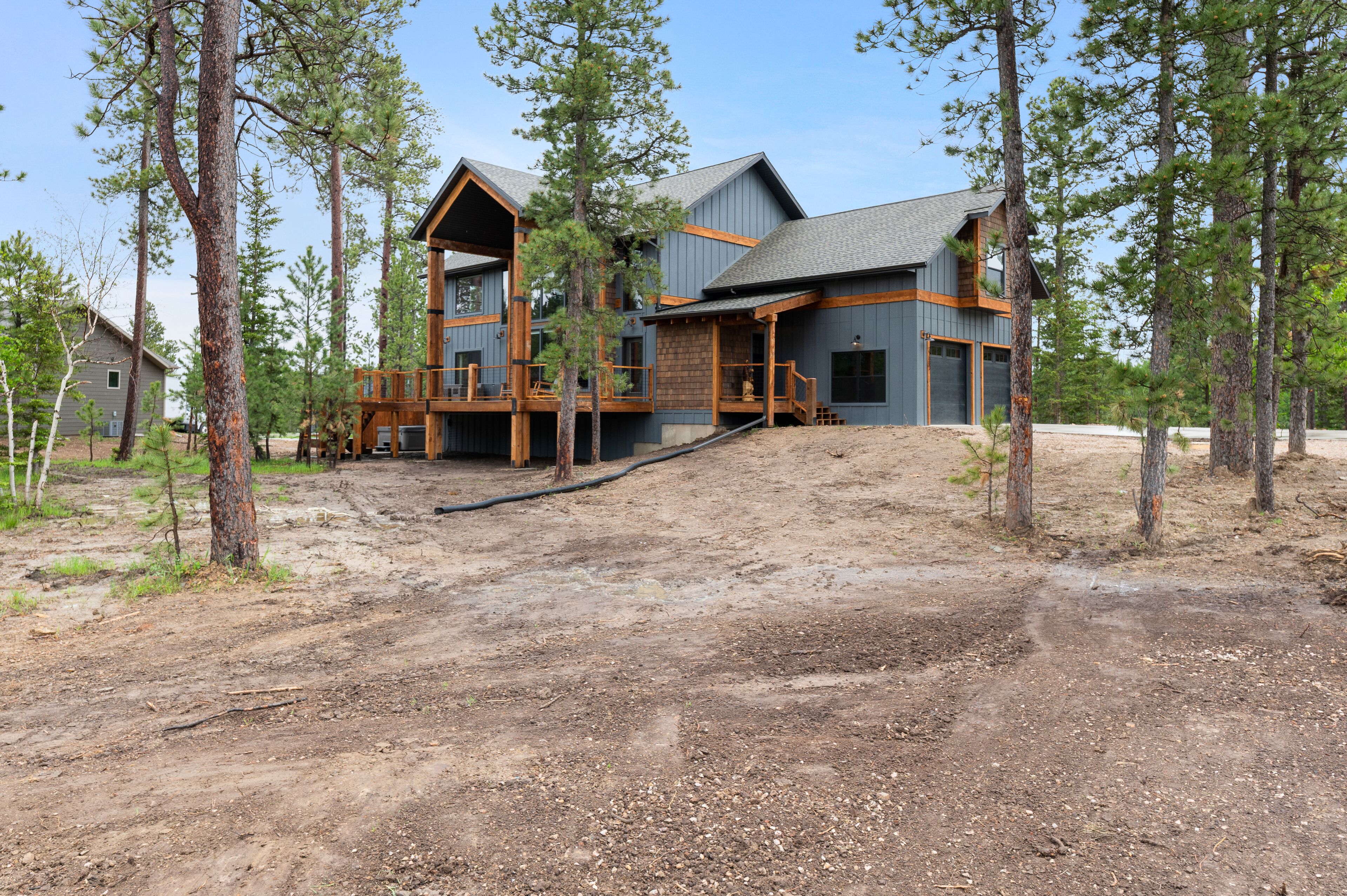 A modern two-story house with a wooden deck and a stone foundation, surrounded by tall pine trees.