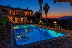 A swimming pool with clear water, surrounded by stone tiles, and a house with a balcony in the background.