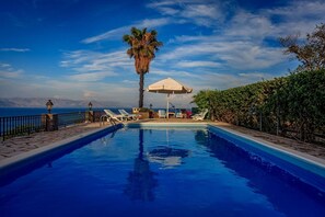 A swimming pool with lounge chairs and an umbrella, surrounded by a stone deck and a view of the sea.