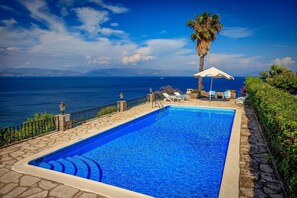 A rectangular swimming pool with a stone border, surrounded by lounge chairs and an umbrella, overlooking a body of water with mountains in the distance.