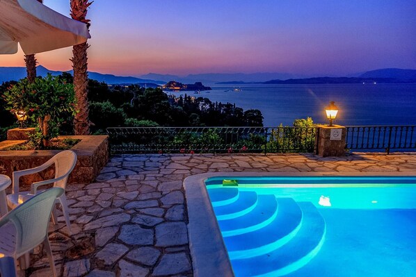 A poolside area with stone flooring, a pool with blue lighting, chairs, and a view of the sea at dusk.