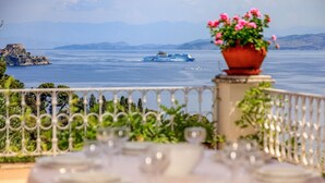 Un balcon d’où l’on aperçoit un navire de croisière au loin, un paysage montagneux et un pot de fleurs orné de fleurs roses.