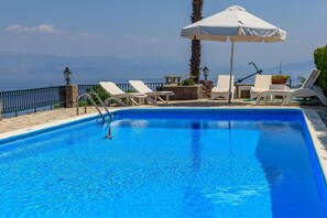 A swimming pool with clear blue water, surrounded by white lounge chairs and a white umbrella.