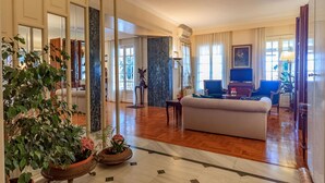 Un grand salon comprenant un canapé, un téléviseur et une table à manger. On y trouve également des plantes en pot et une colonne en marbre.