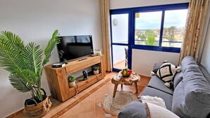 Un salon avec un téléviseur posé sur un meuble en bois, un canapé gris, une petite table ronde sur laquelle se trouve un saladier rempli de fruits, une grande plante en pot et une vue sur l’extérieur par une fenêtre à cadre bleu.