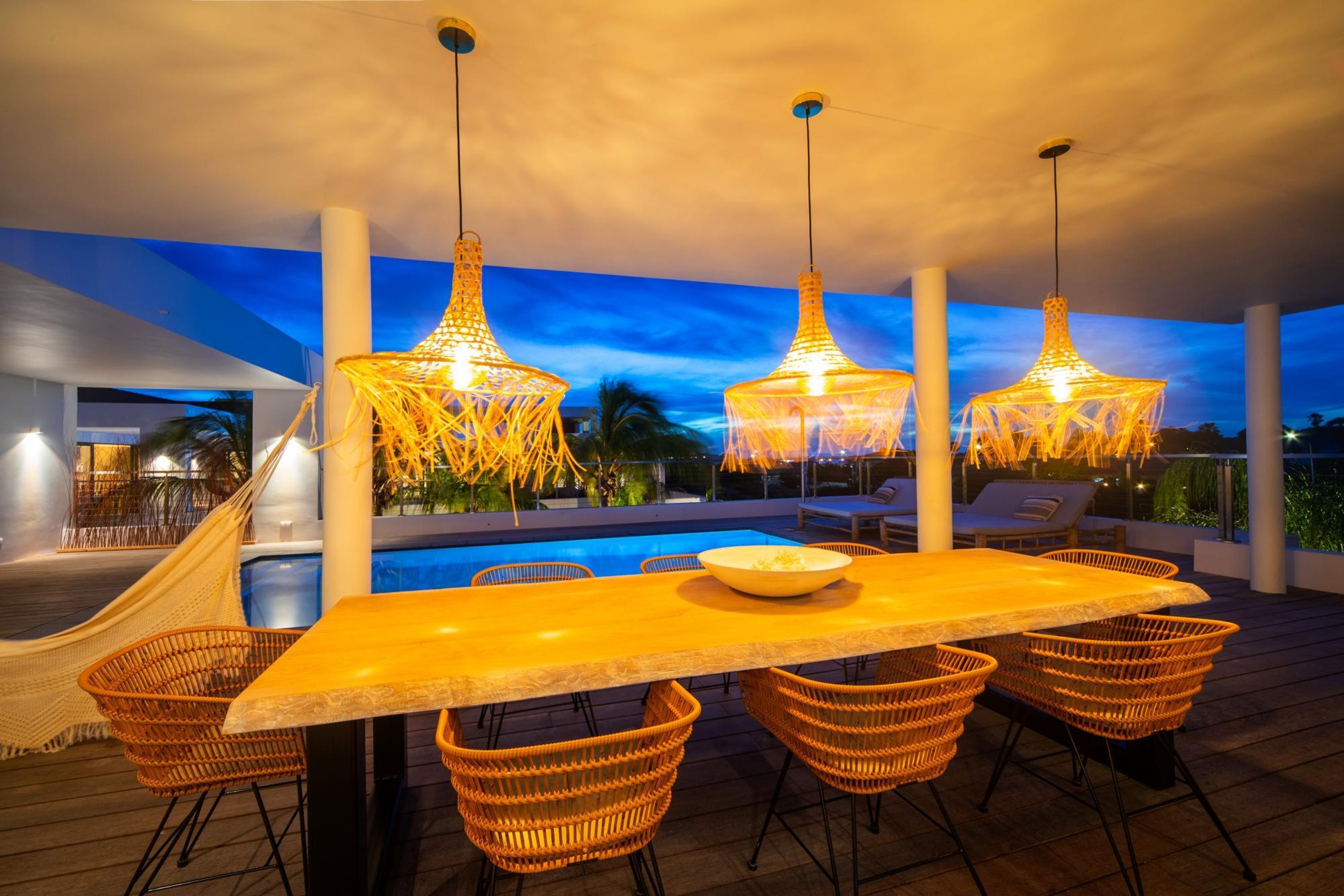 An outdoor dining area with a pool, wicker chairs, and hanging lights.