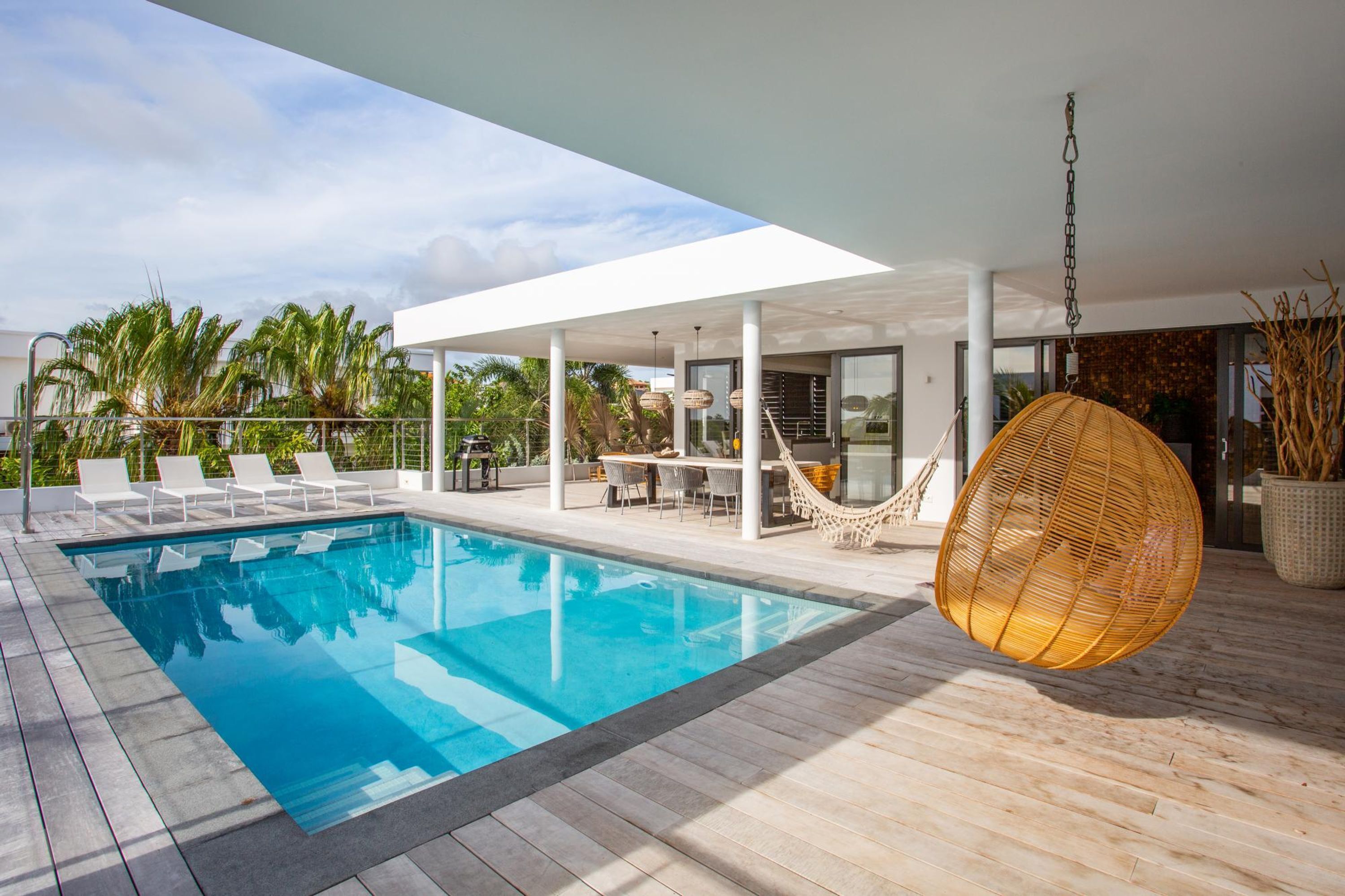 Un área de piscina moderna al aire libre, con una piscina azul cristalina, sillas de descanso blancas, una silla colgante y un espacio de estar cubierto con una mesa y sillas.