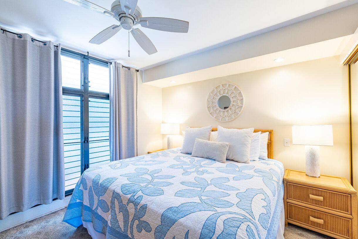 Een hotelkamer met een bed, twee bedlampen, een plafondventilator, een groot raam met gordijnen en een spiegel aan de muur.