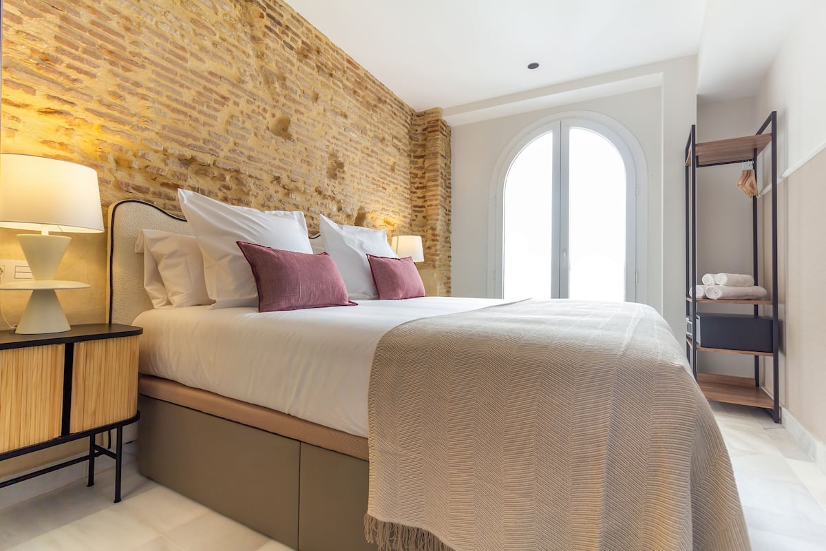 Ein Schlafzimmer mit Backsteinwand, einem Bett mit weißer und pinkfarbener Bettwäsche, einem Nachttisch mit Lampe und einem Kleiderschrank mit Spiegel.