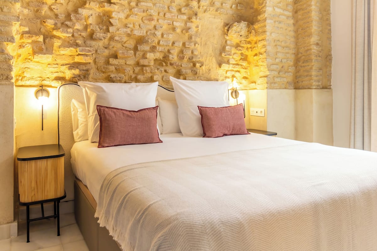 Ein Hotelzimmer mit Backsteinwand, einem Bett mit weißer Bettwäsche und zwei rosafarbenen Kissen, einem Nachttisch mit Lampe und einem Beistelltisch.