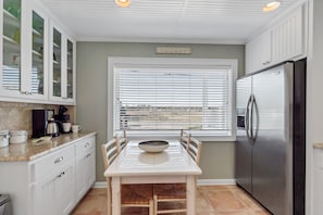 A kitchen with white cabinets, a stainless steel refrigerator, a dining table with chairs, and a window with blinds.
