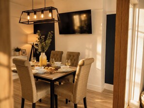A dining area with a wooden table, four upholstered chairs, a vase with greenery, a bowl of grapes, and a bottle of wine. - Family Modern 3 Bedroom hot tub House (Blackpool)