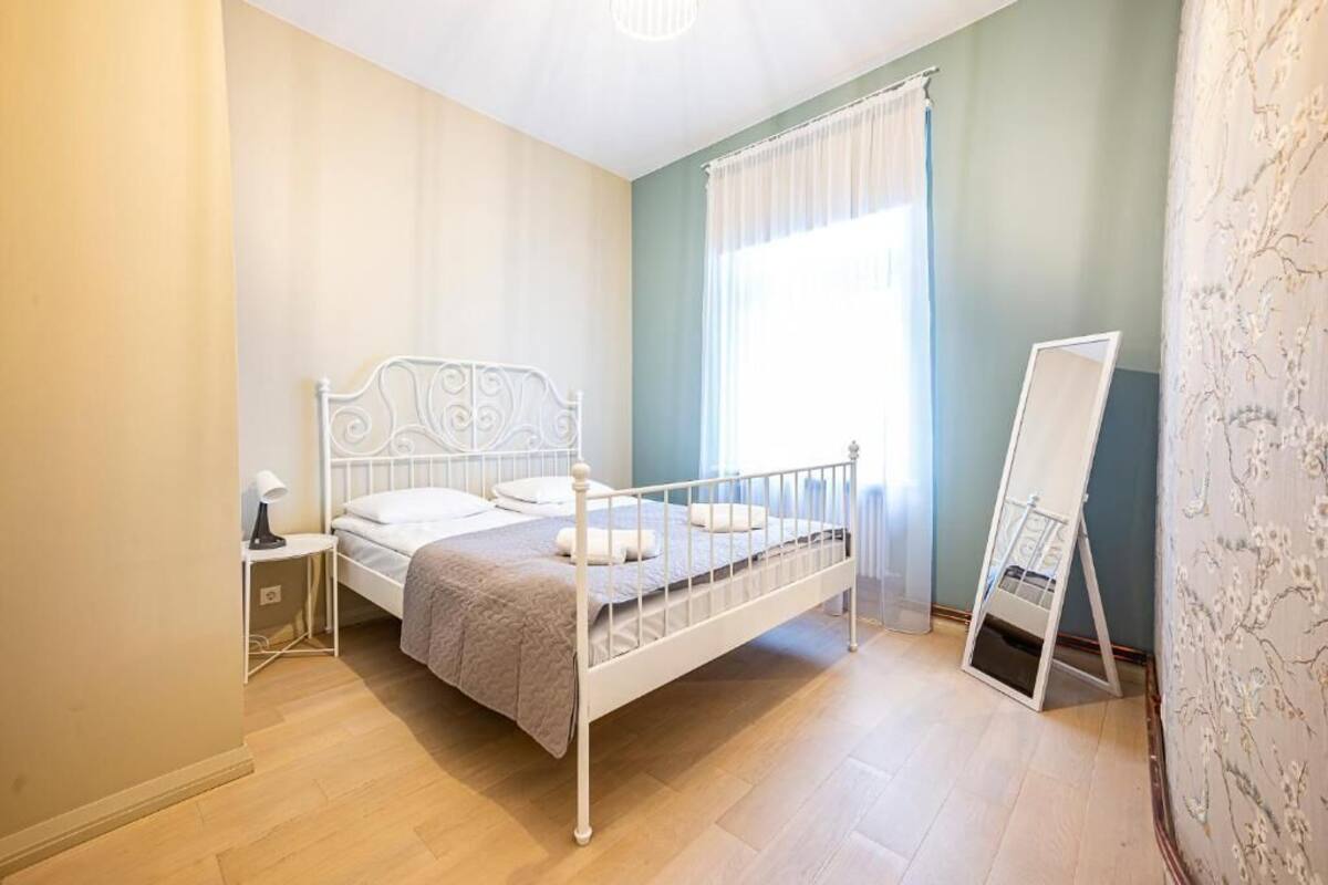 Un dormitorio con una estructura de cama de metal blanco, una mesita de noche con lámpara, una ventana con cortinas y un papel tapiz con diseño.