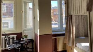 Une pièce avec un plafond haut, des moulures décoratives, une grande fenêtre donnant sur des immeubles, un radiateur, un lit à baldaquin et une chaise.