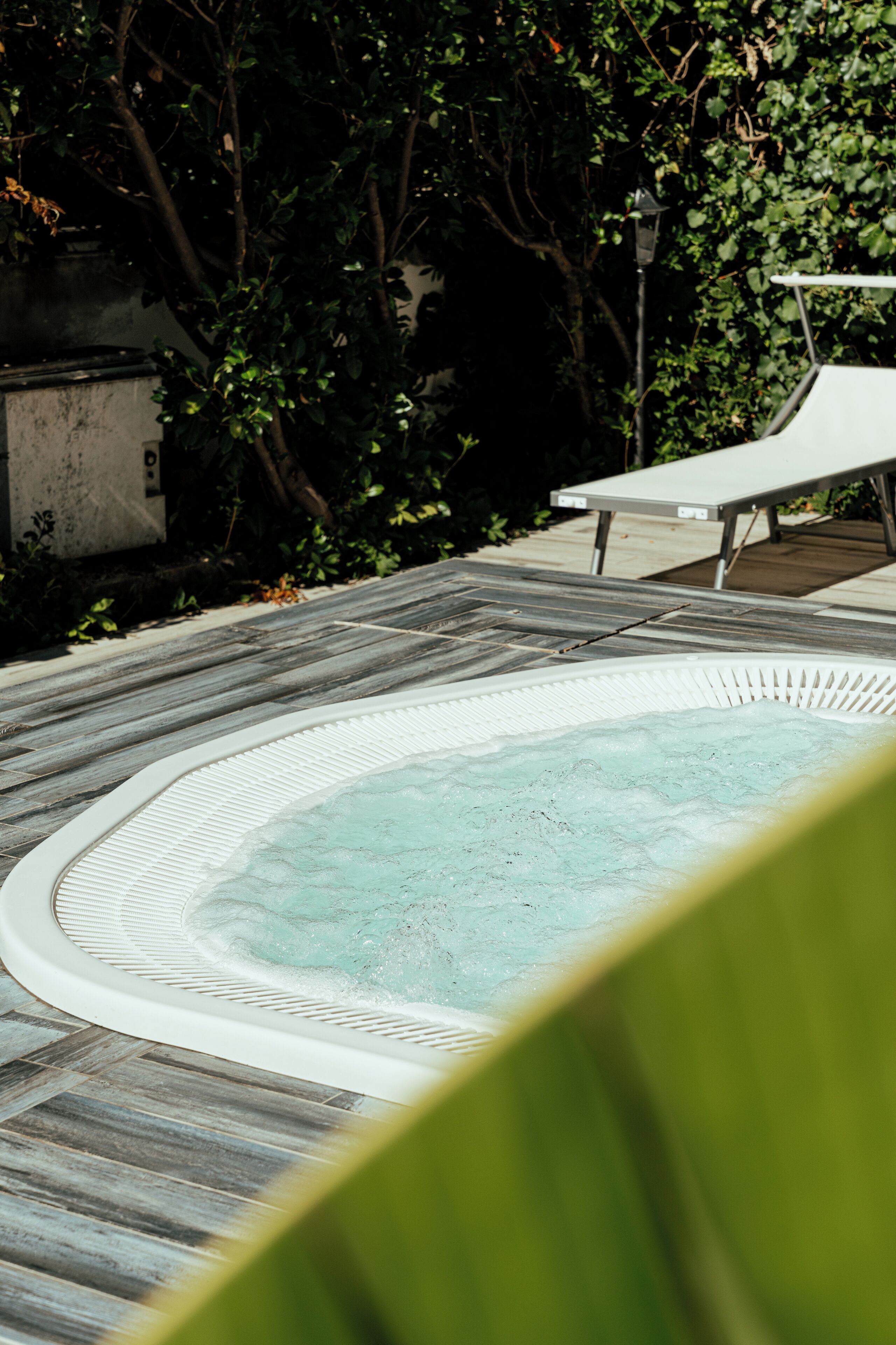 Una bañera de hidromasaje rodeada por una terraza de madera y follaje, con una silla de descanso cerca.