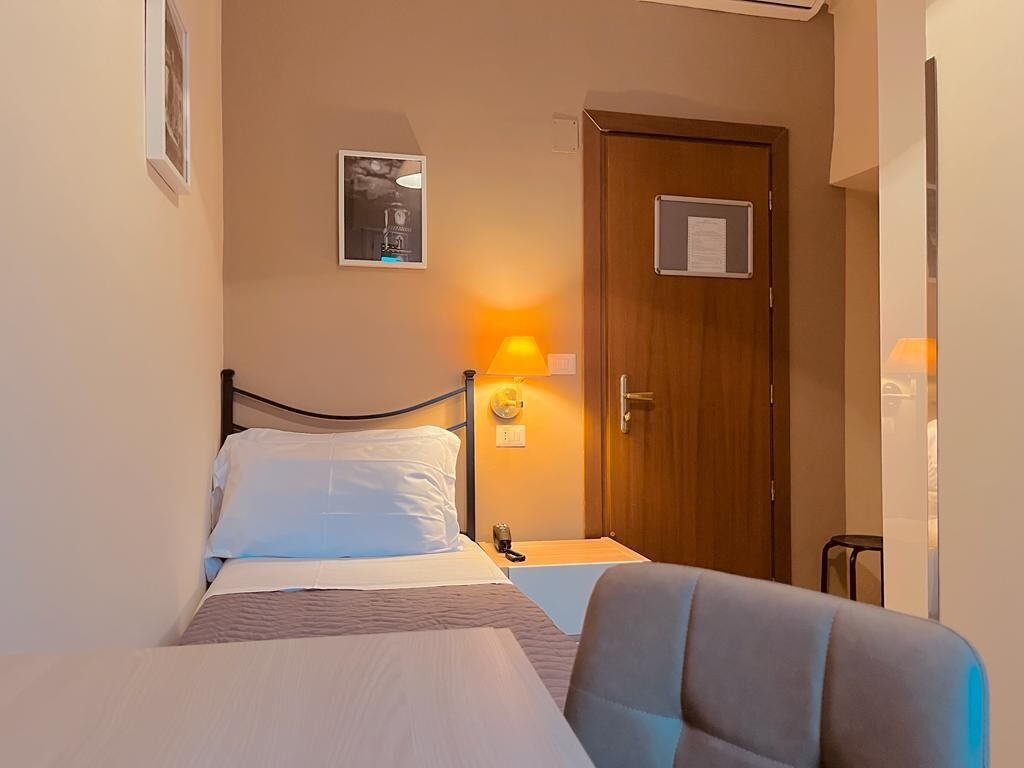 A hotel room with a bed, a bedside table with a lamp, a wooden door, and a small chair.