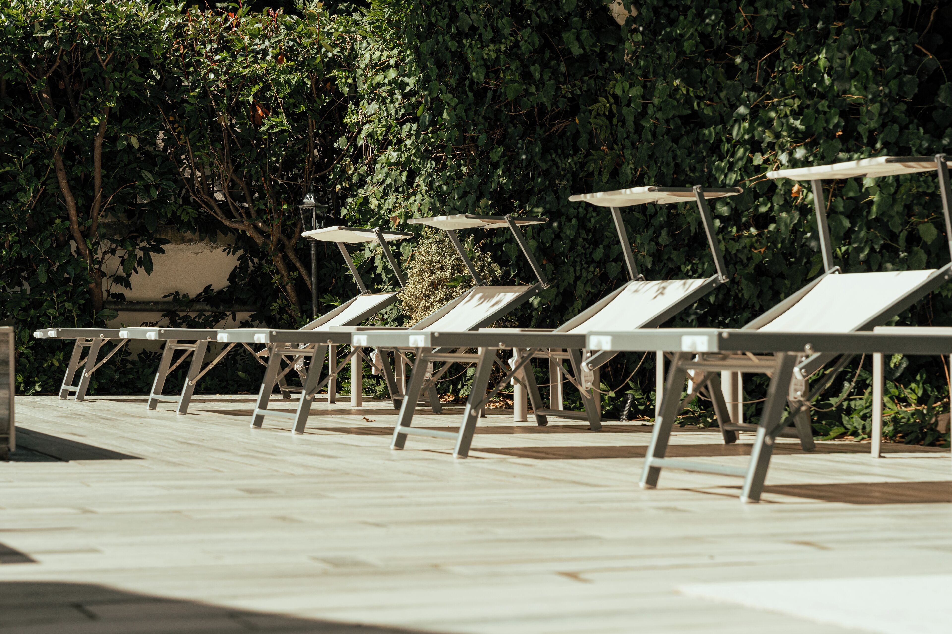 A row of sun loungers with metal frames and white cushions, aligned on a tiled patio area with a backdrop of dense green foliage.