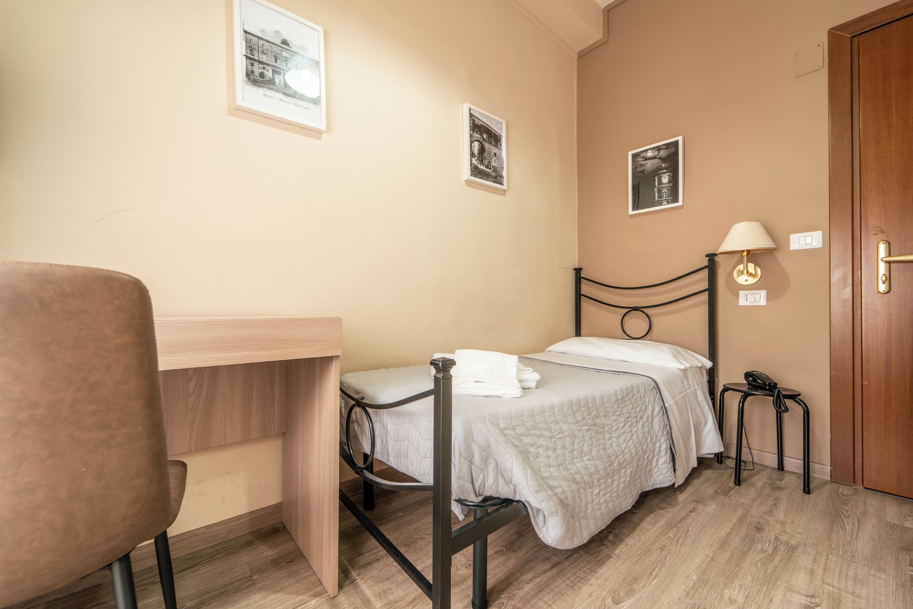 Een kleine, netjes ingerichte slaapkamer met een enkel bed, een bureau, een stoel en een nachtkastje met een lamp.