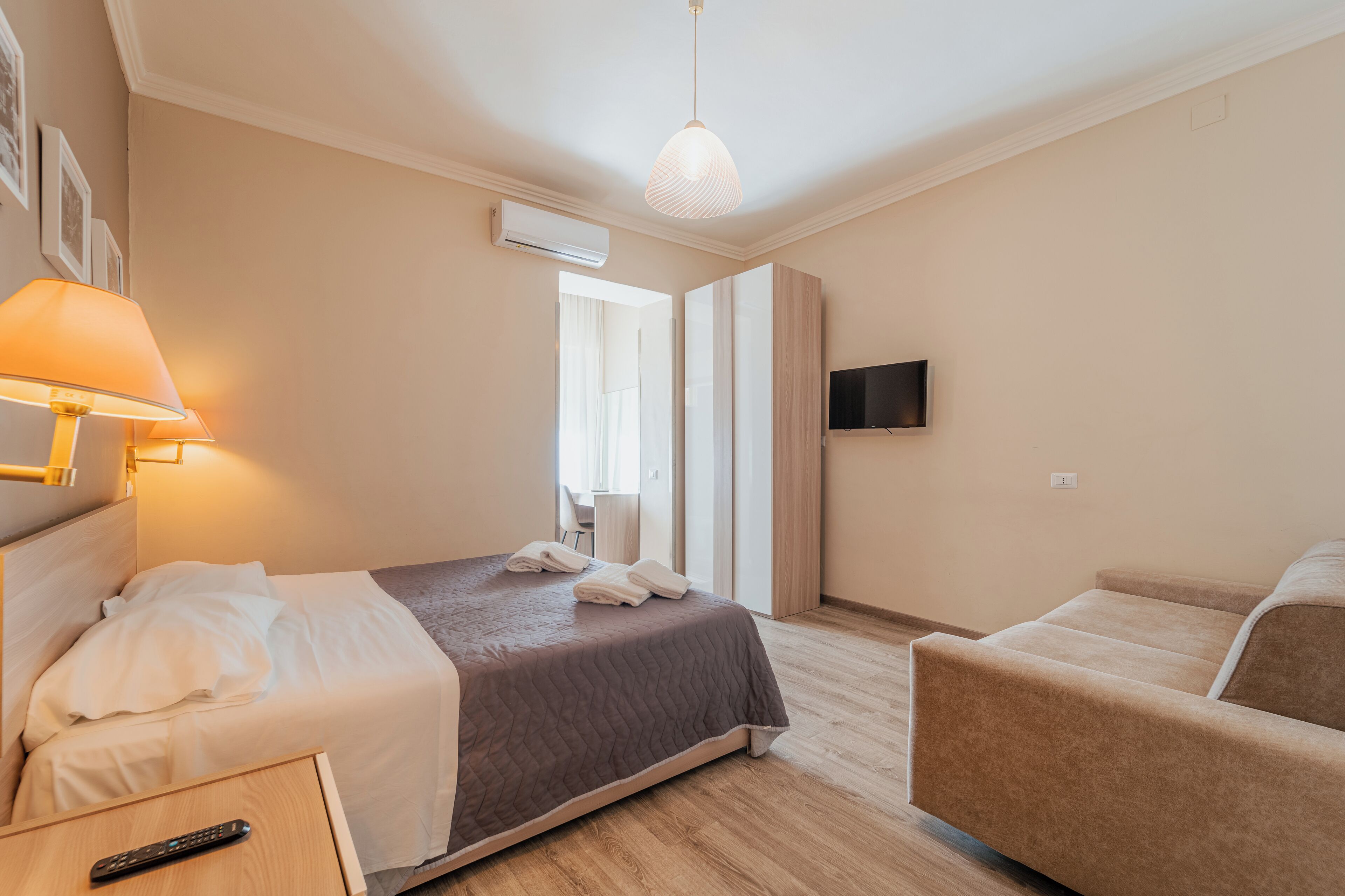 A hotel room with a bed, a bedside table with a remote control, a sofa, a TV mounted on the wall, and a hanging light fixture.