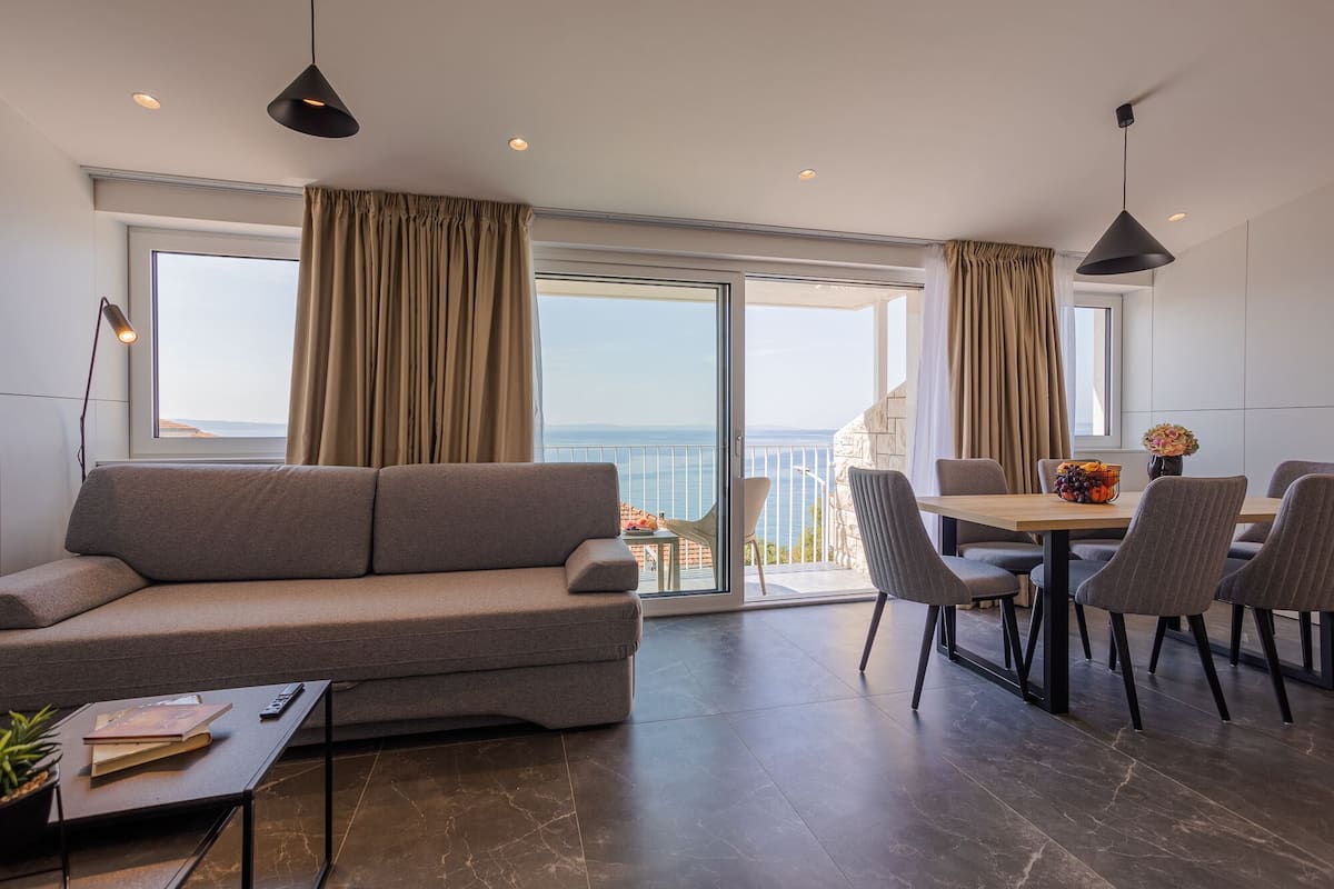 A modern living room with a sofa, dining table, and chairs, a balcony with a view of the sea, and a potted plant on a side table.