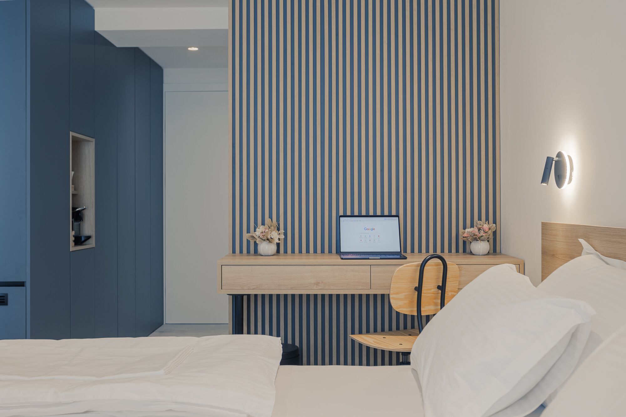 Une chambre d’hôtel moderne équipée d’un lit, d’un bureau avec un ordinateur portable, d’une chaise et d’une table de chevet agrémentée d’un vase de fleurs.