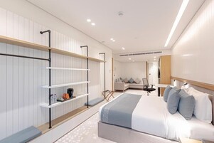 Una habitación de hotel moderna con una cama grande, una zona de estar con sofá y sillones, y estanterías empotradas con espacio de almacenamiento.