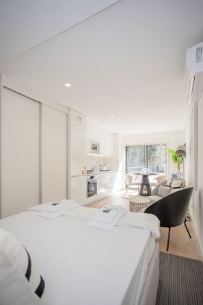 Camera d'albergo moderna con un letto grande, un'area pranzo con tavolo e sedie, un angolo cottura e un'ampia finestra che lascia entrare luce naturale.